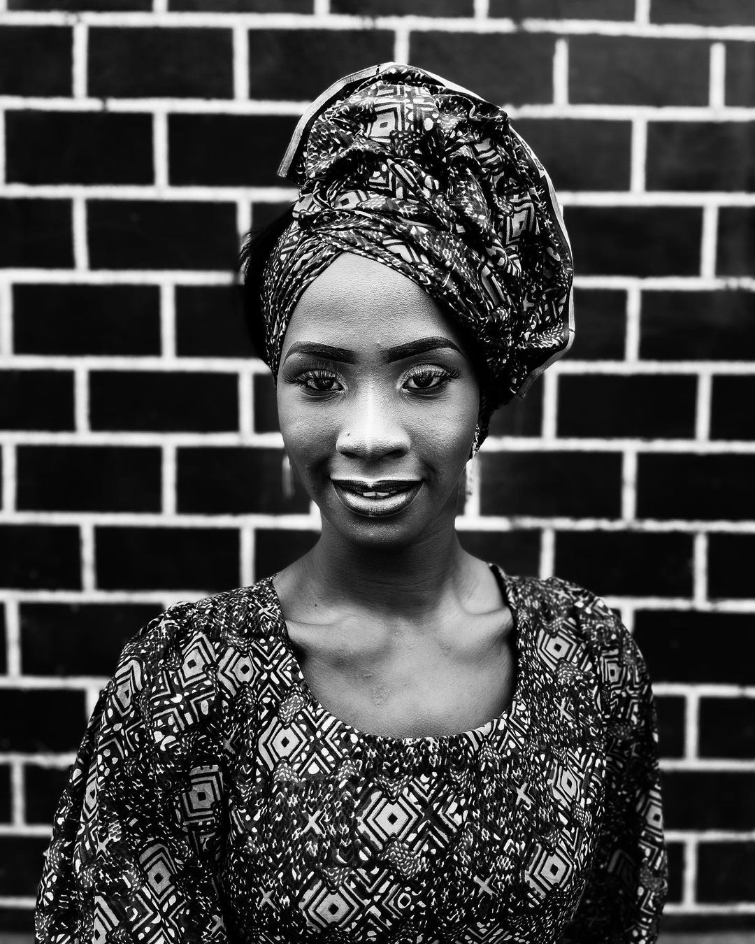 Portrait of an adult female with a patterned headwrap and top posing in front of a dark brick wall in a close-up studio shot.