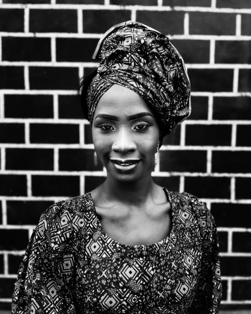 Portrait of an adult female with a patterned headwrap and top posing in front of a dark brick wall in a close-up studio shot.