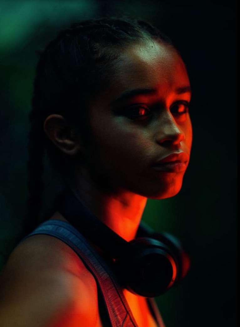 Profile portrait of an adult woman with braided hair wearing headphones around her neck, illuminated with red and green lighting.