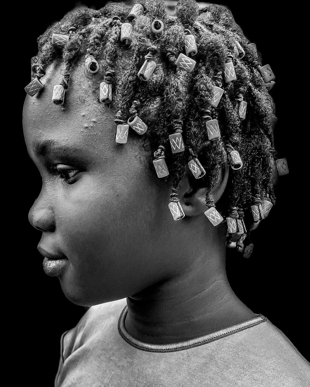 Monochrome portrait of a child with decorative beads in braided hair, looking to the side against a dark background, close-up.