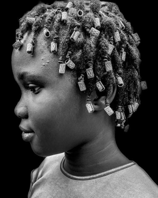 Monochrome portrait of a child with decorative beads in braided hair, looking to the side against a dark background, close-up.