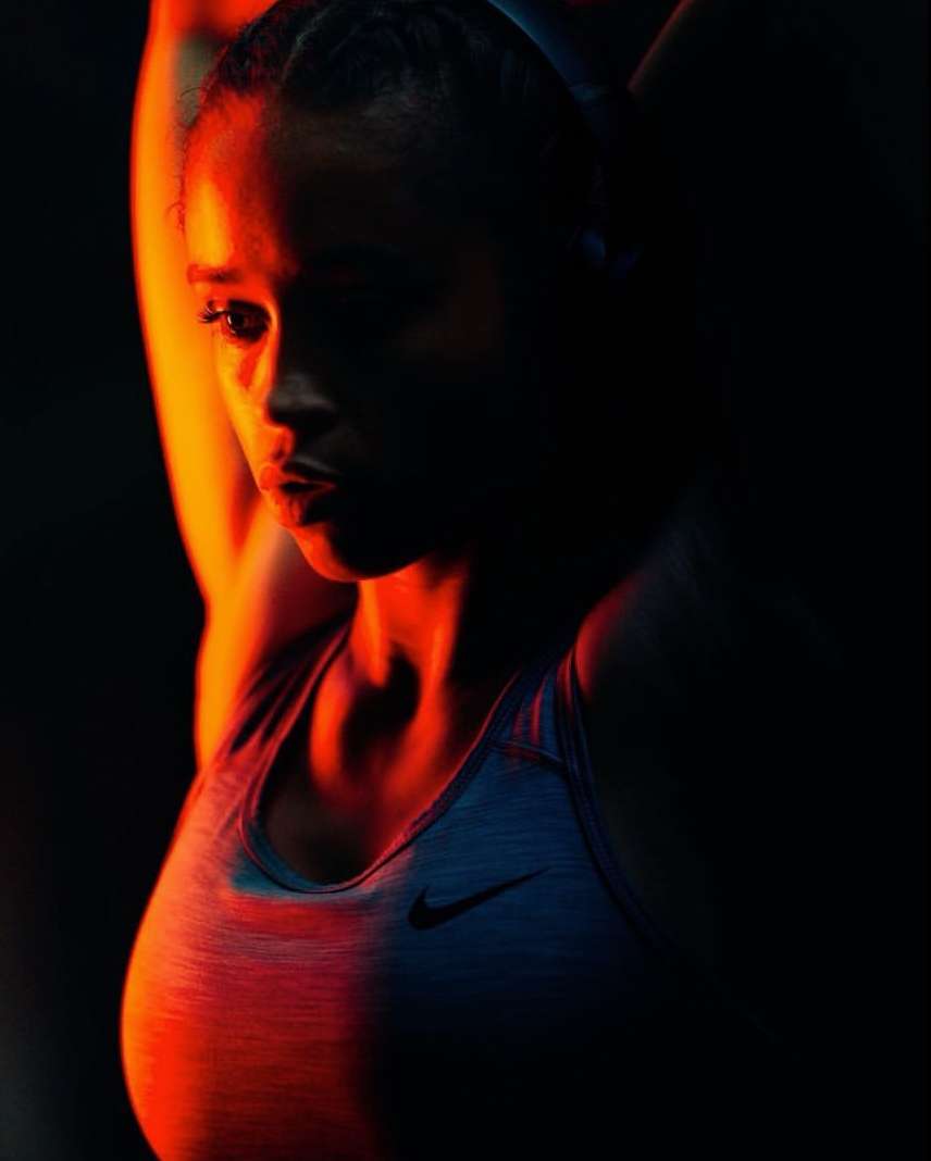 Athletic woman in sports bra poses with arms raised in a dimly lit studio with orange and red lighting for a fitness portrait.