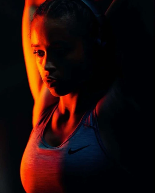 Athletic woman in sports bra poses with arms raised in a dimly lit studio with orange and red lighting for a fitness portrait.