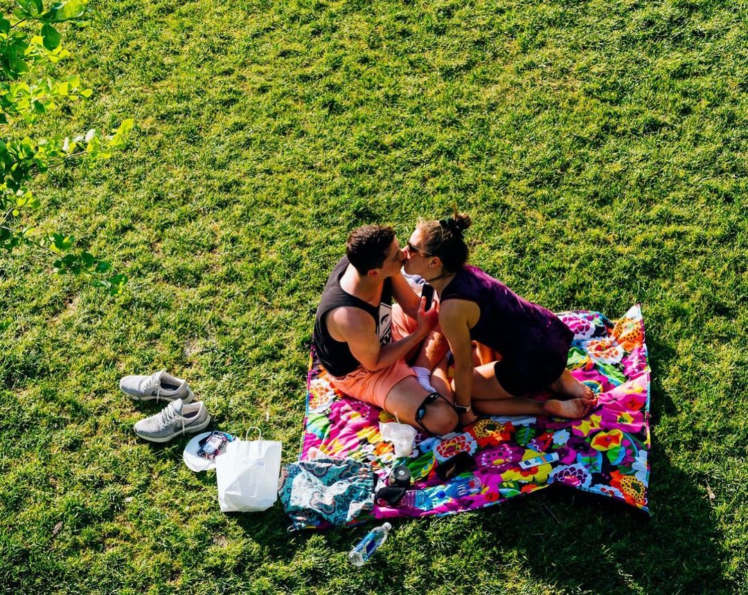 Couple sharing a kiss on a colorful blanket in a sunlit grassy park, enjoying a sunny day with shoes and personal items nearby.