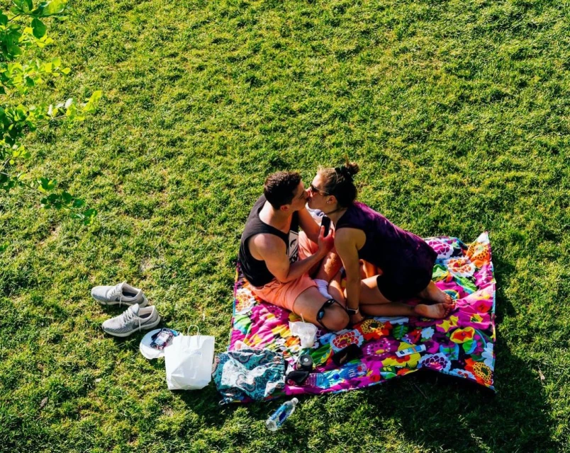 Couple sharing a kiss on a colorful blanket in a sunlit grassy park, enjoying a sunny day with shoes and personal items nearby.