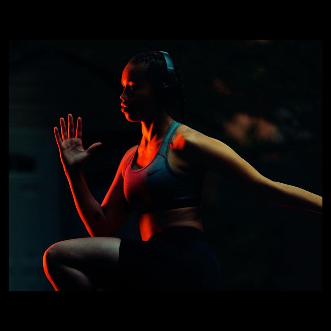 Athletic woman in sports bra and shorts, wearing headphones, is exercising in a dark, shadowed environment with red accent lighting.