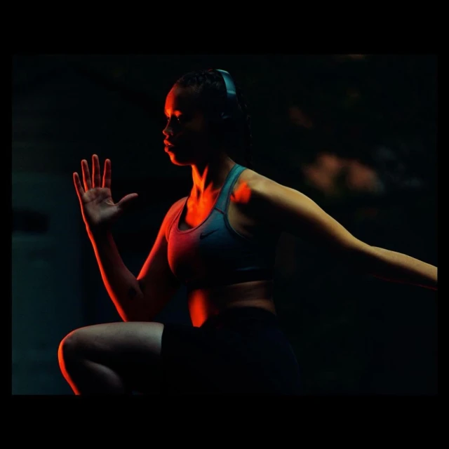 Athletic woman in sports bra and shorts, wearing headphones, is exercising in a dark, shadowed environment with red accent lighting.