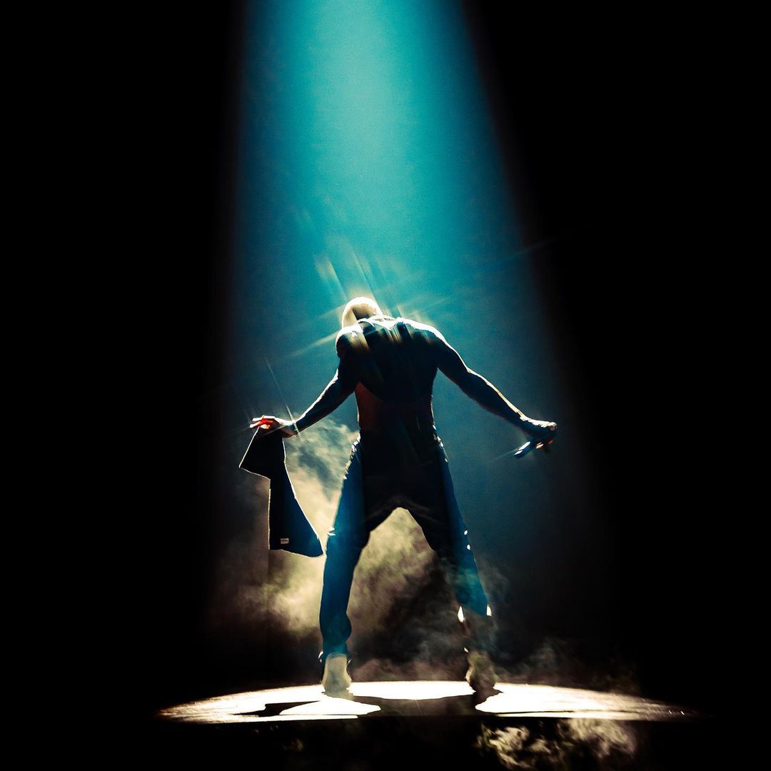 Male dancer under a bright blue spotlight, holding a garment, in front of a dark background with smoke, performing live.