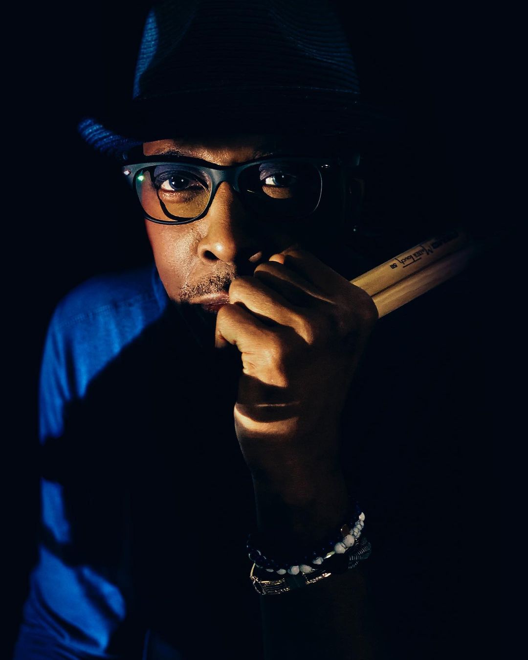Portrait of a Black adult male wearing glasses and a hat, holding drumsticks in a dark indoor setting with bright spotlight.