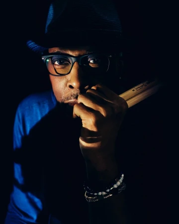 Portrait of a Black adult male wearing glasses and a hat, holding drumsticks in a dark indoor setting with bright spotlight.