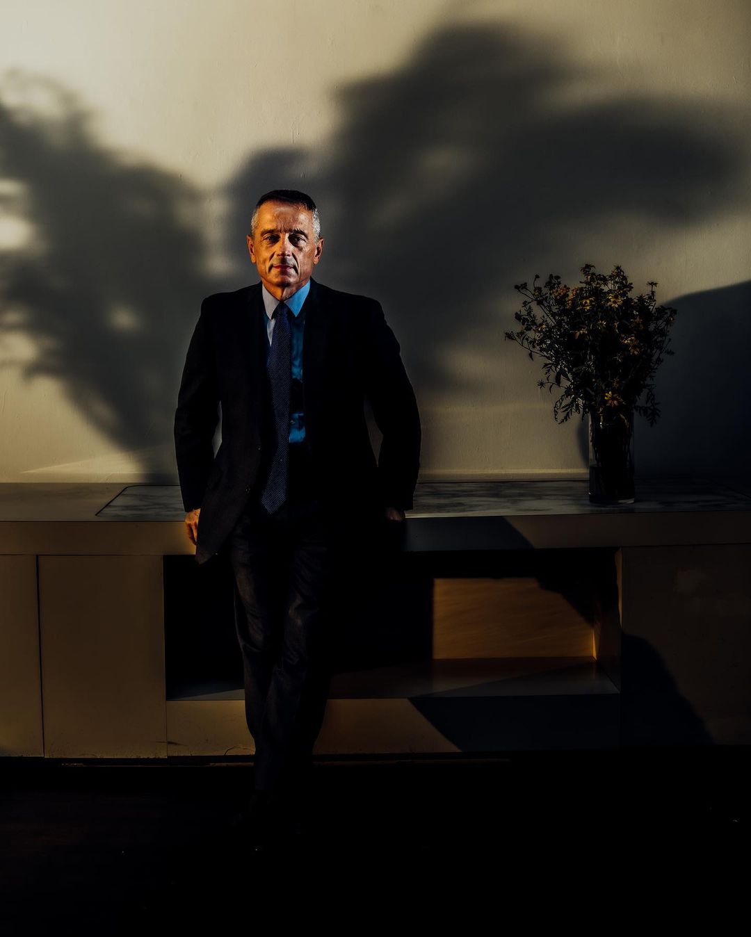 Portrait of a middle-aged man in a suit indoors, posed against a cream wall with shadow patterns and a cabinet holding flowers in a vase.
