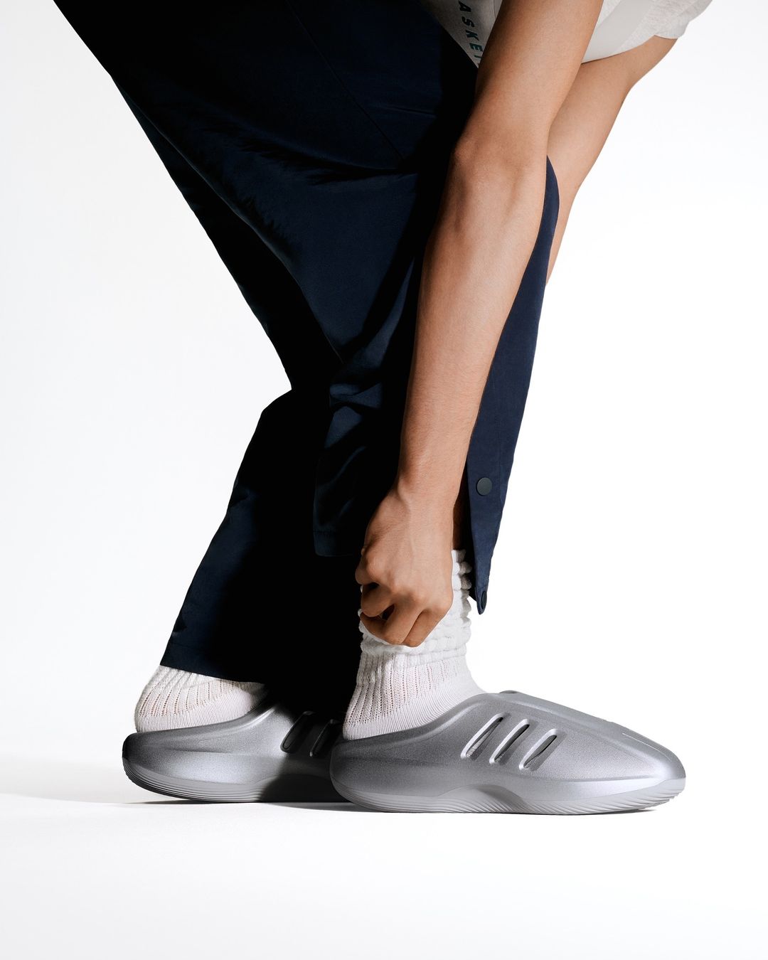 Person in dark pants and grey socks adjusts the cuff of their pants, wearing slip-on style shoes on a bright white backdrop.