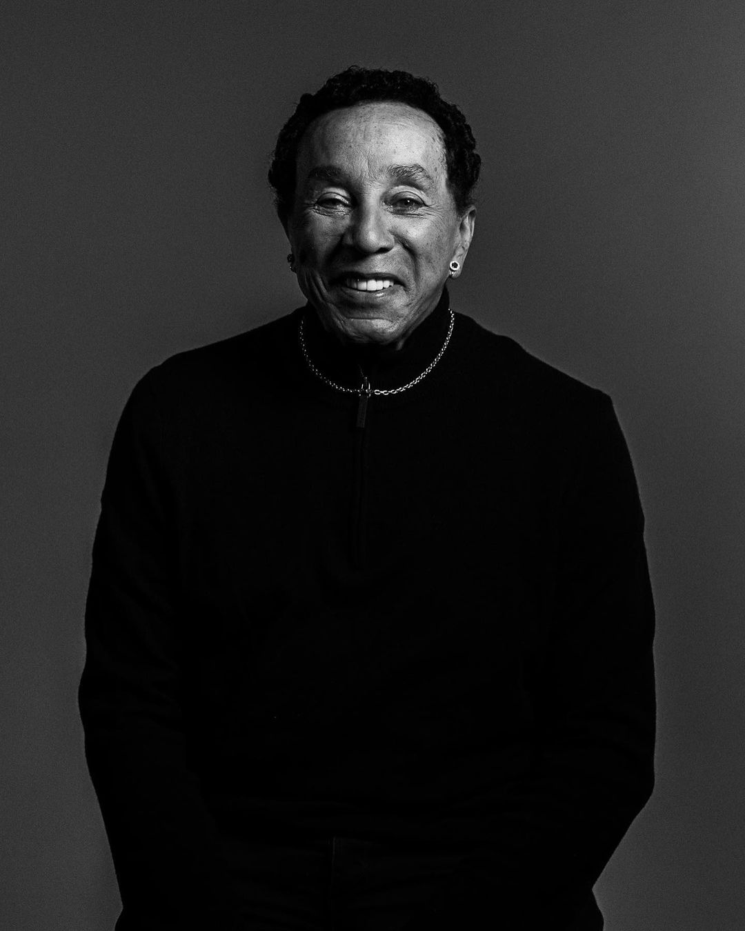 Black and white portrait of an adult man with dark hair smiling, wearing a black jacket and a silver necklace on a gray backdrop.