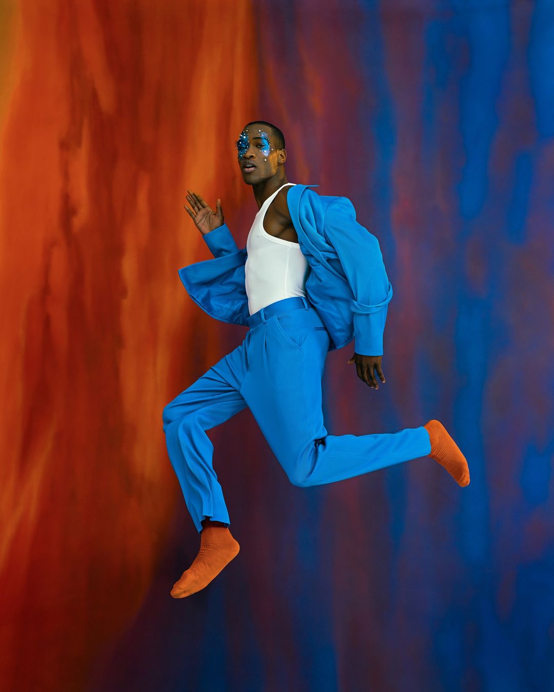Man in blue suit and orange socks leaps against a colorful background, showcasing fashion and movement in a dynamic studio portrait.