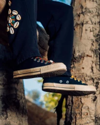 Close-up shot of person wearing black trousers with cowrie shell embellishments and black sneakers with white rubber toes outdoors against a tree.