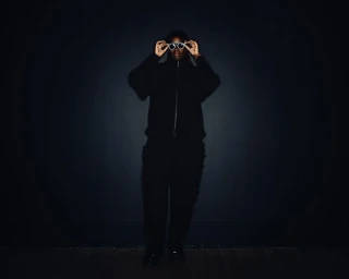 Person wearing all black clothing holding up round sunglasses in a dimly lit studio setting