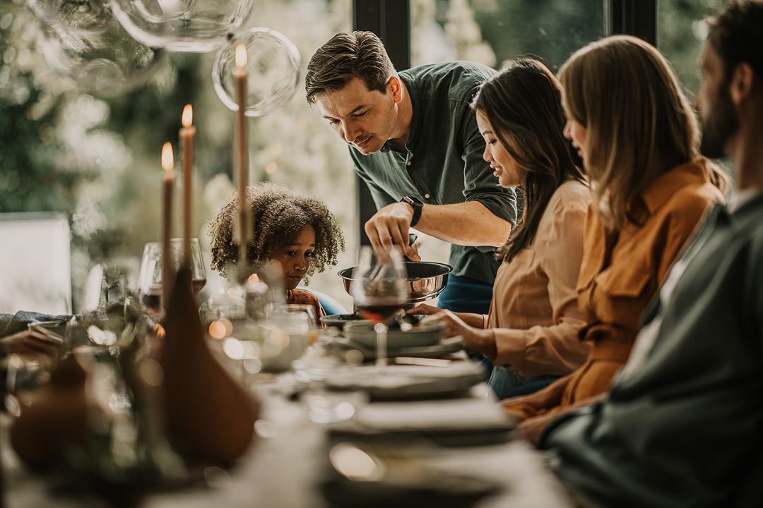 A family gathers at a dinner table with candles in an indoor setting, featuring a man serving food from a pan and a child looking on.