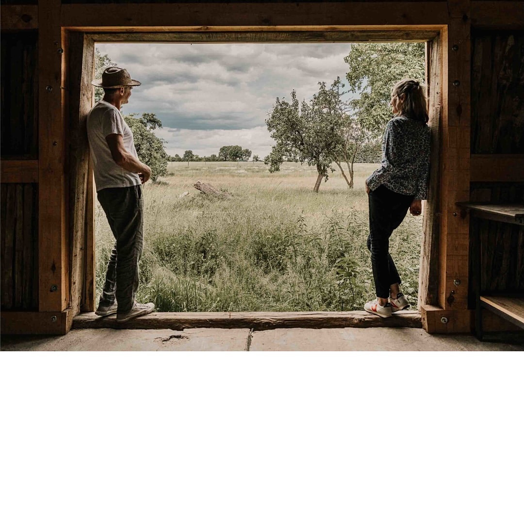 An adult couple stands in the opening of a rustic wooden structure, looking out over a grassy field under a cloudy sky in daylight.