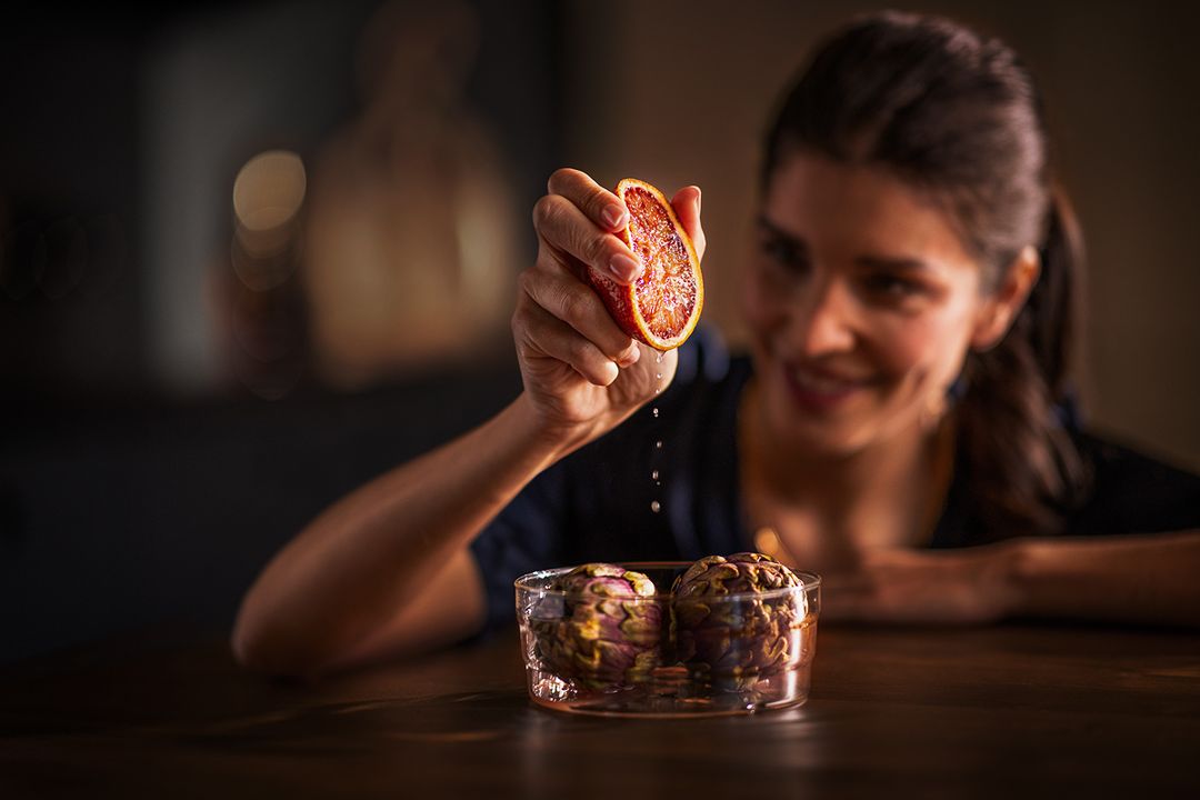 Woman squeezes blood orange into glass jar with artichokes on wooden table in warm indoor lighting, focus on the fresh juice.