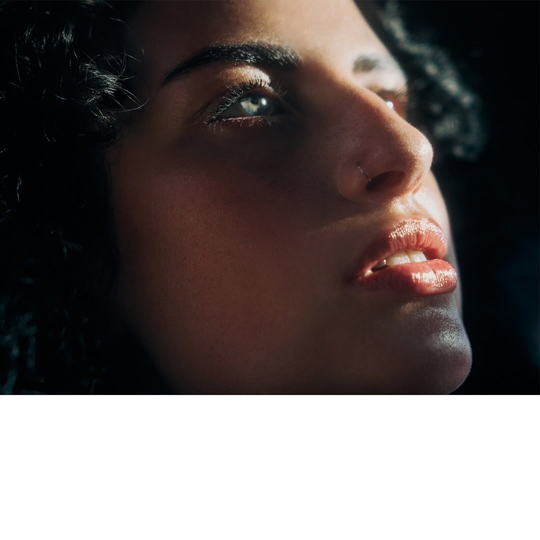 Adult female portrait showcasing skin, nose ring, and curly dark hair, captured with an eye-level, medium shot, set against a dark background.