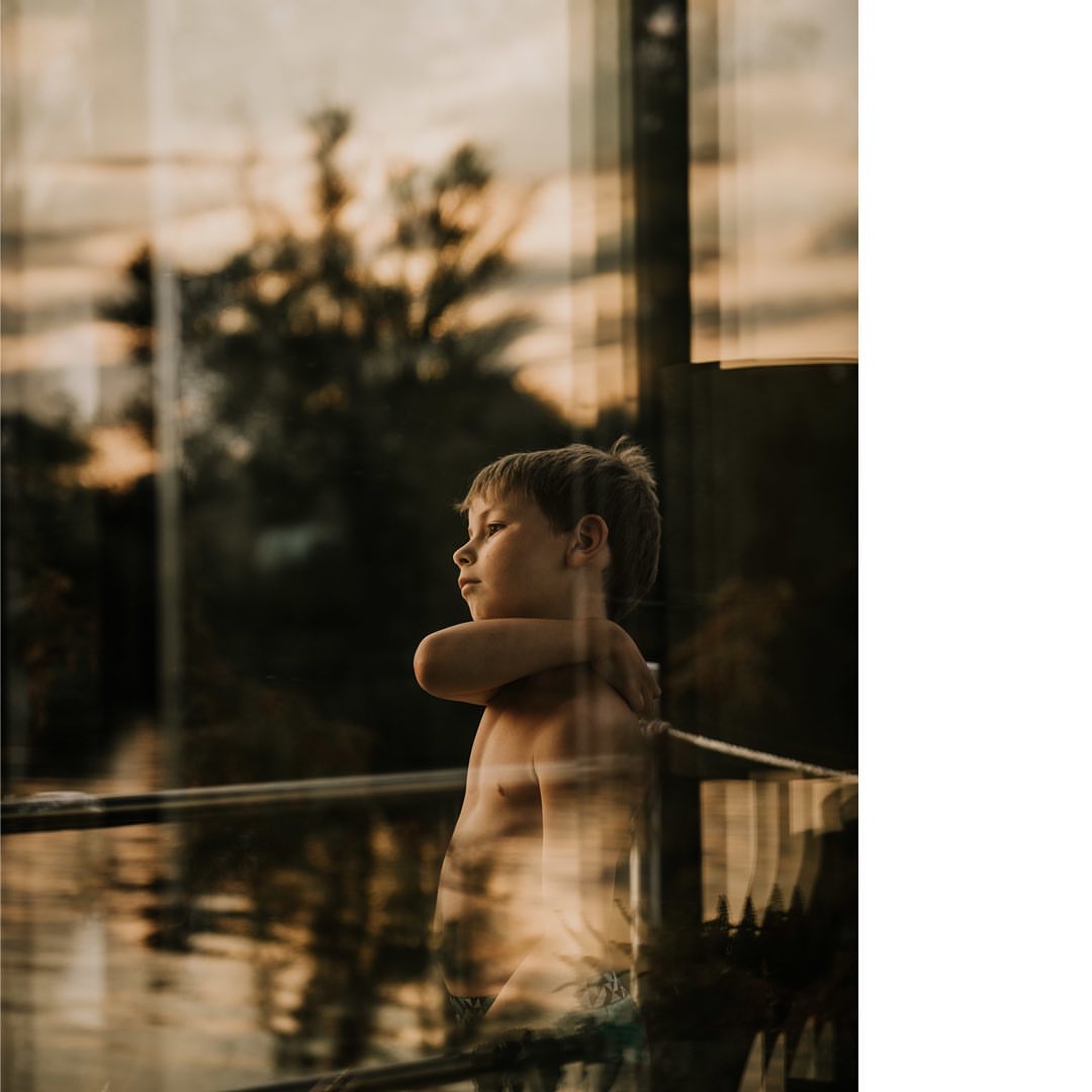 Young boy standing indoors reflected in a window, looking out toward trees and sky at dusk, arms crossed, pensive mood.