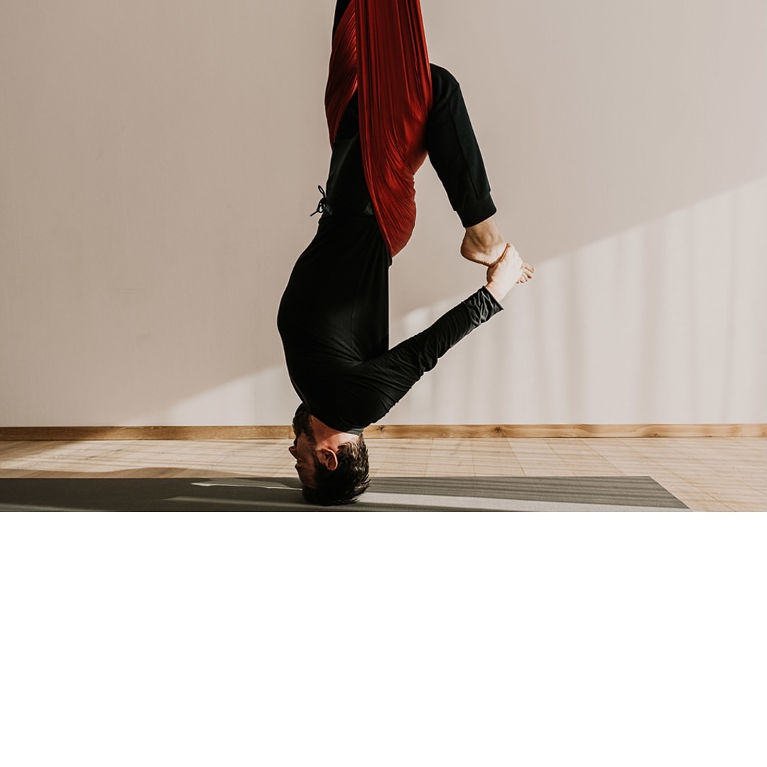 Adult male in black performing inverted yoga pose with red aerial silk in a studio with wooden floor and natural lighting.