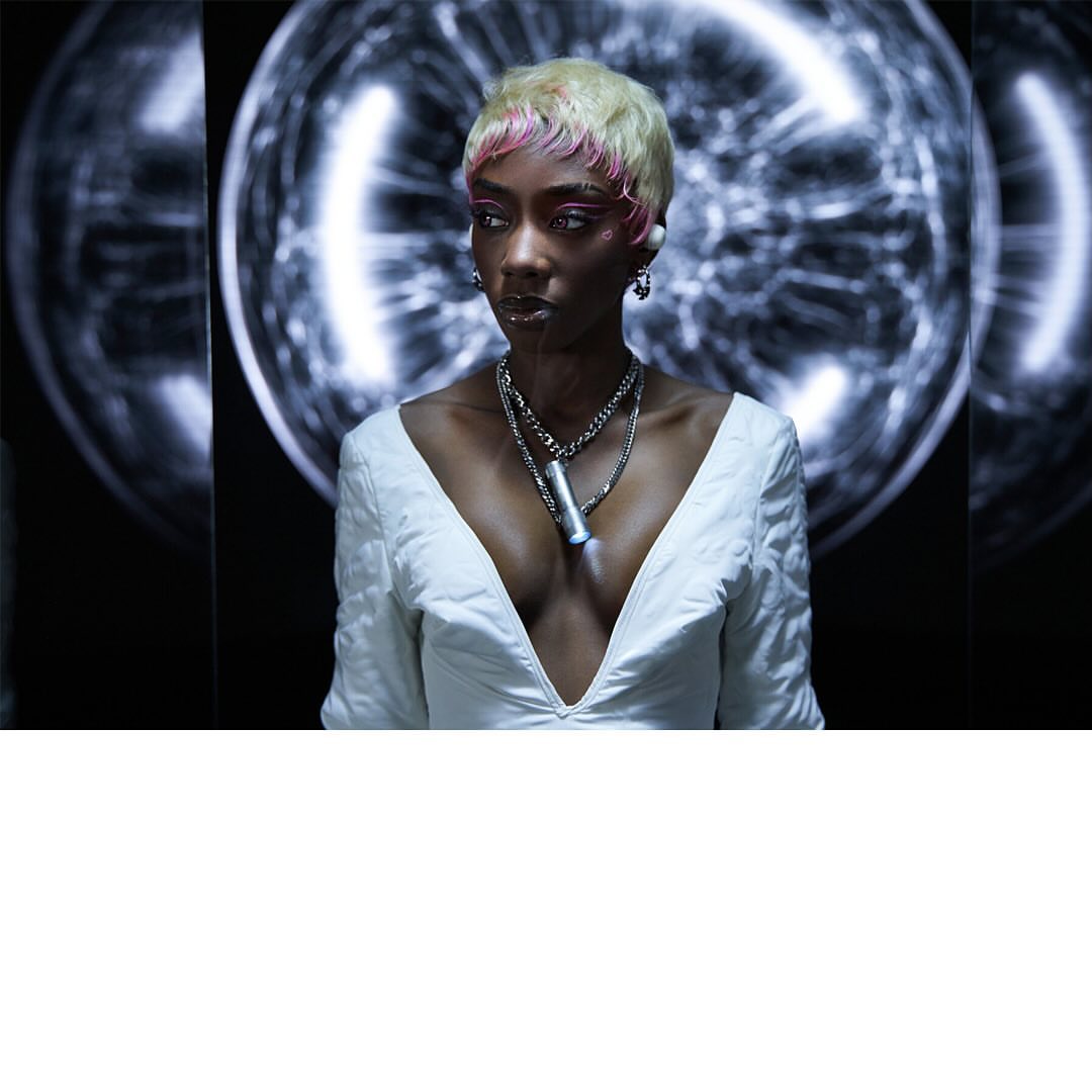 Portrait of a person with short blond hair and pink roots wearing a white dress and silver necklaces, posing in a dark interior.
