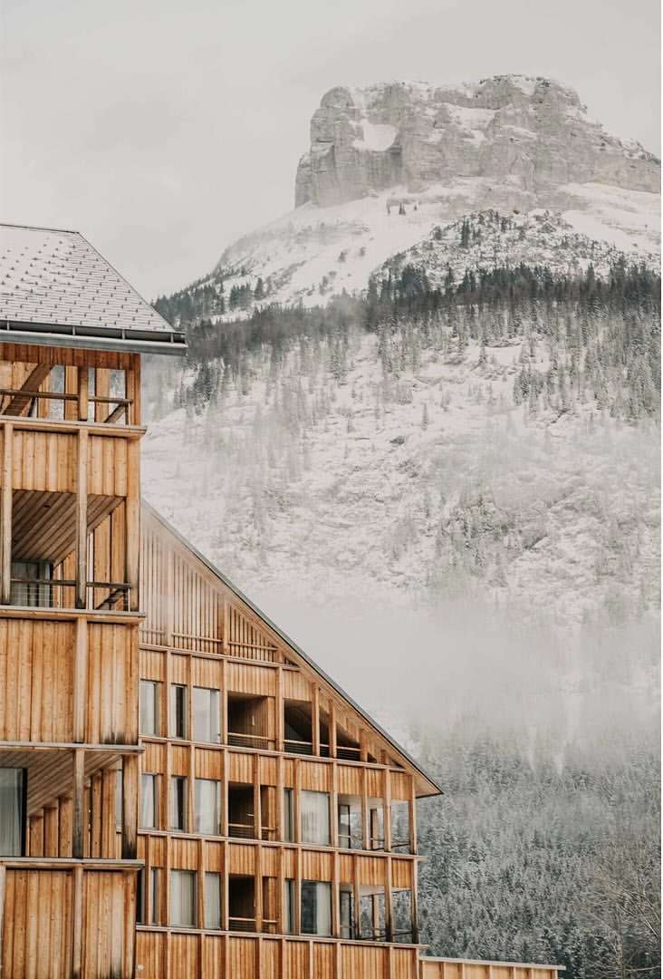 Exterior of a wooden hotel with balconies, set against a backdrop of snow-covered mountains and a dense forest under an overcast sky.