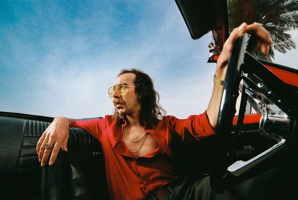 Adult male in red shirt and aviator glasses driving a black convertible car under a sunny blue sky with palm trees. 
