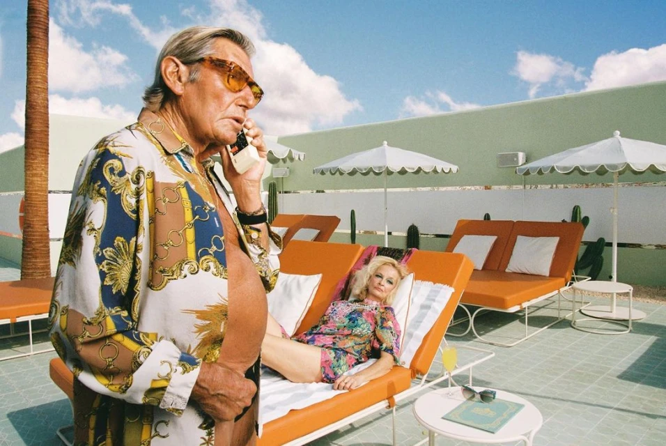 Older man in patterned shirt talking on retro phone, woman relaxing on lounger by pool on a sunny day; leisure and summer vibes.