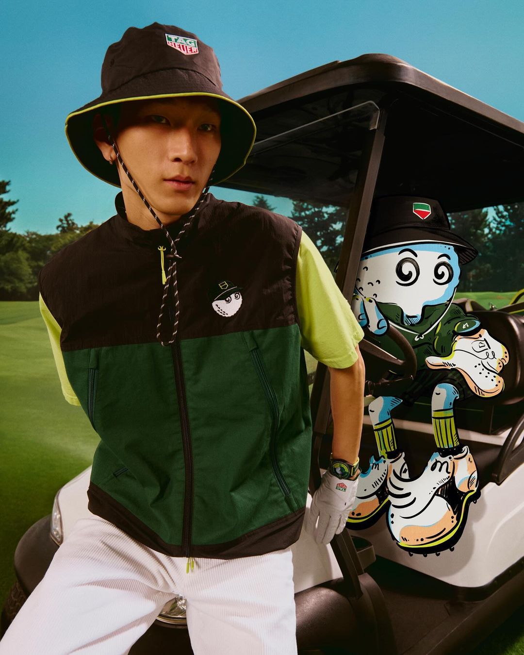 Asian man in golf attire poses with a cartoon golf-themed character riding in a golf cart on a sunny golf course.