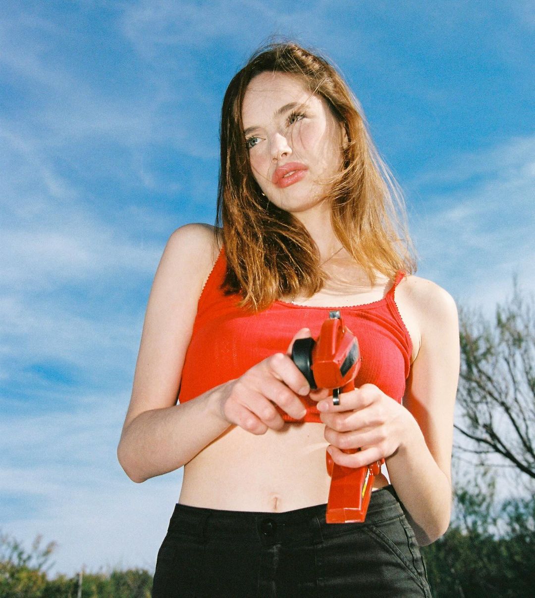 Low angle shot of a young adult woman in a red top holding a red and black toy in an outdoor setting with a blue sky backdrop.