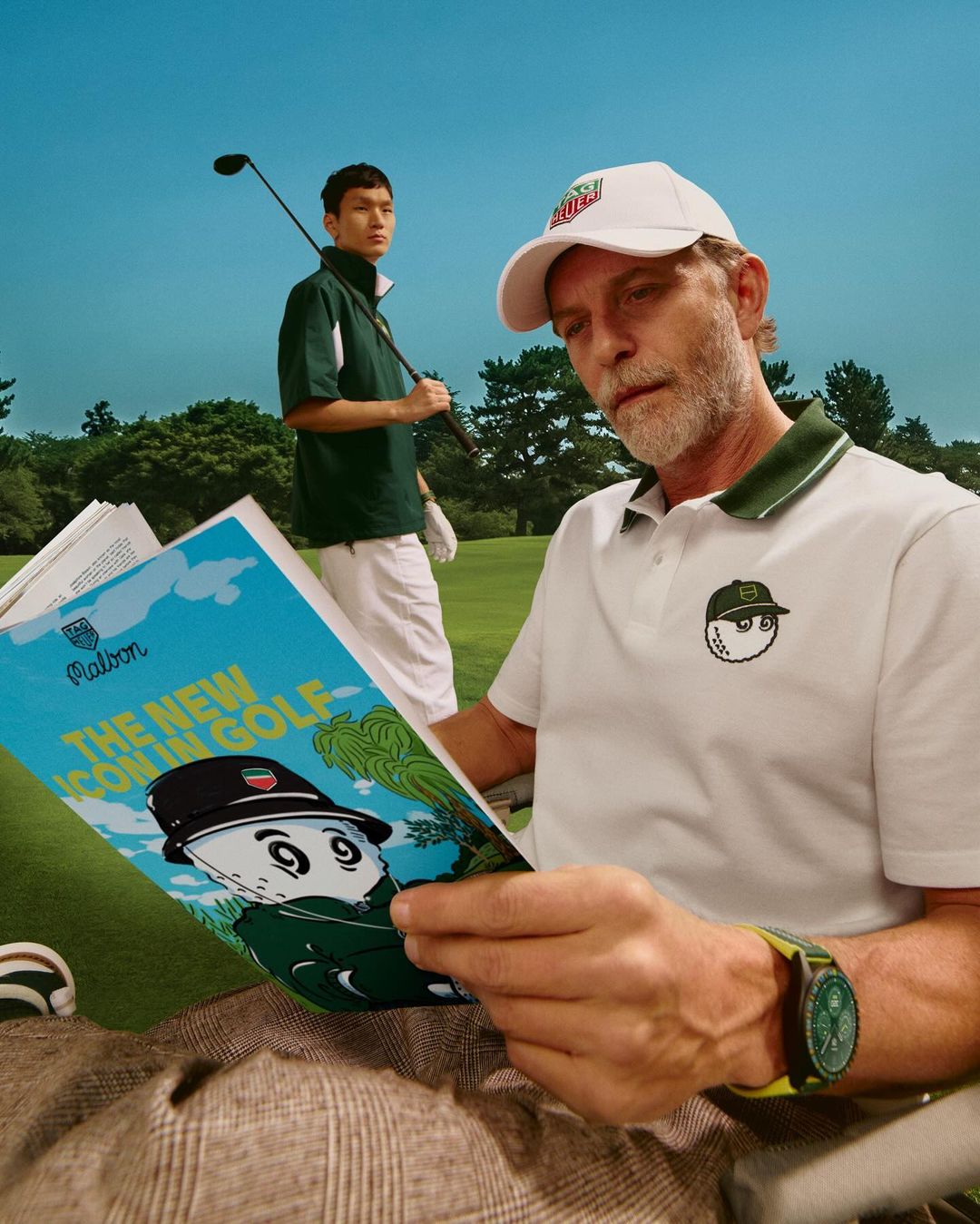 Two men on a golf course, one reading a golf magazine, wearing Malbon apparel; the other carrying a golf club in the background.