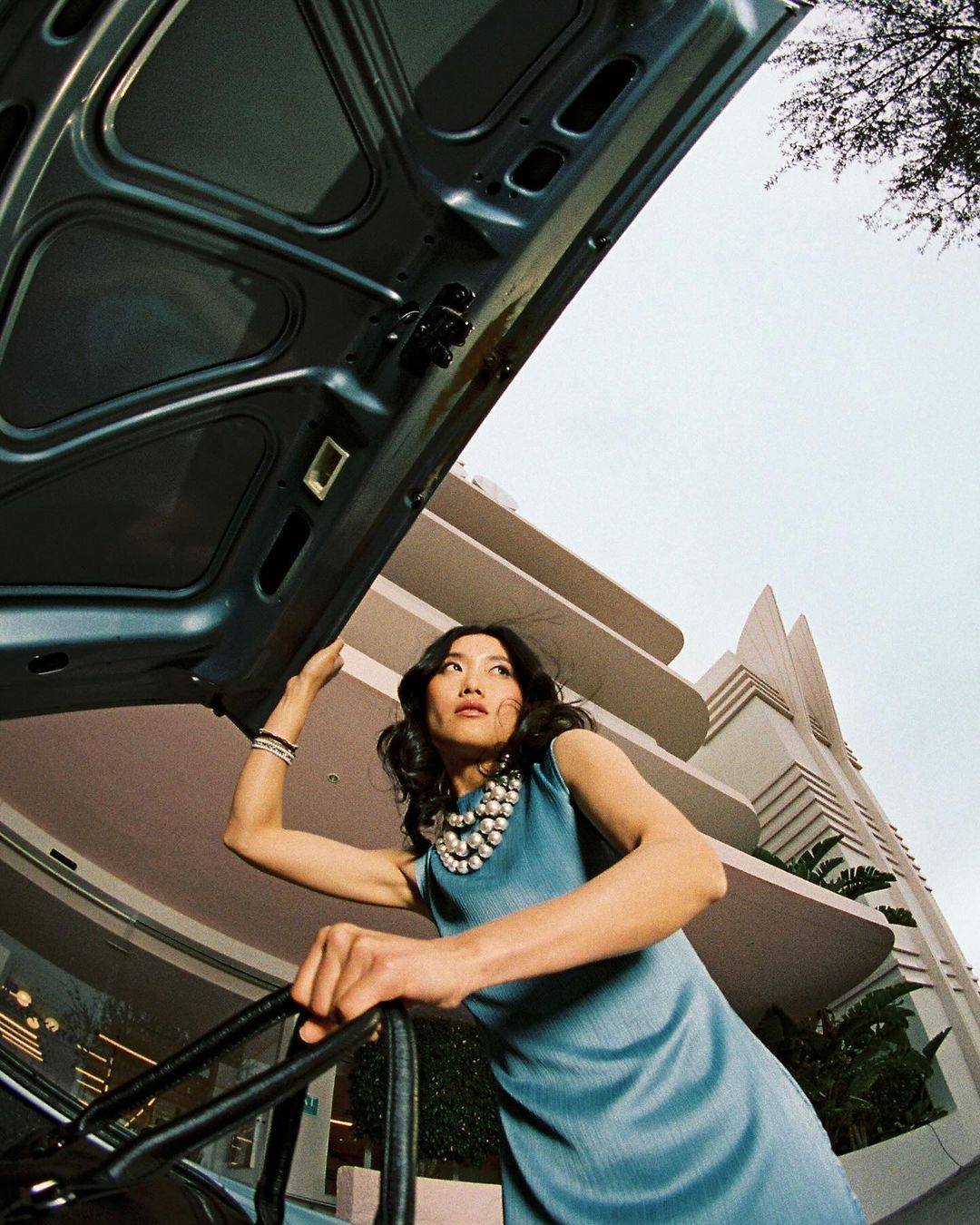 Woman in blue dress stands next to open car hood outside art deco building. Low angle shot capturing fashion and architecture.