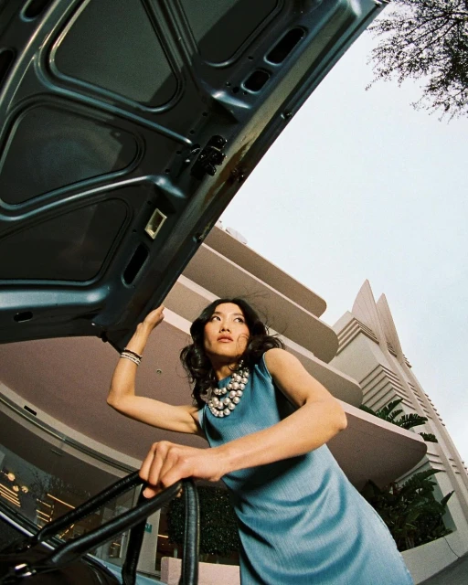 Woman in blue dress stands next to open car hood outside art deco building. Low angle shot capturing fashion and architecture.