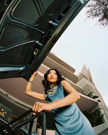 Woman in blue dress stands next to open car hood outside art deco building. Low angle shot capturing fashion and architecture.