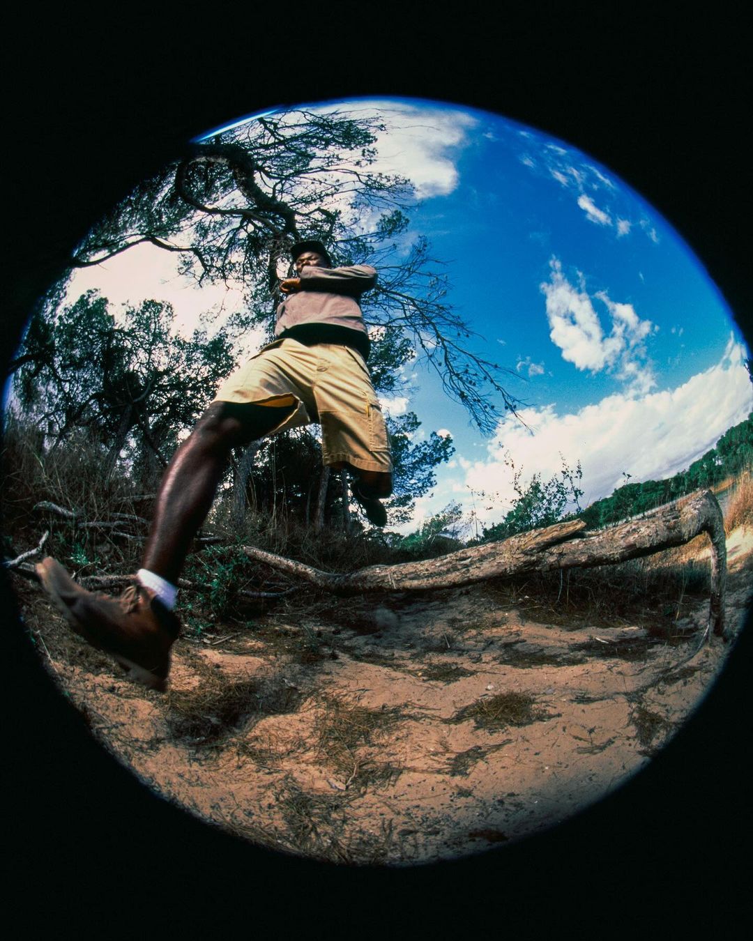Man in mid-air wearing tan shorts and brown shoes. Fisheye lens shows trees, sky, and clouds with a black background around the circular image.