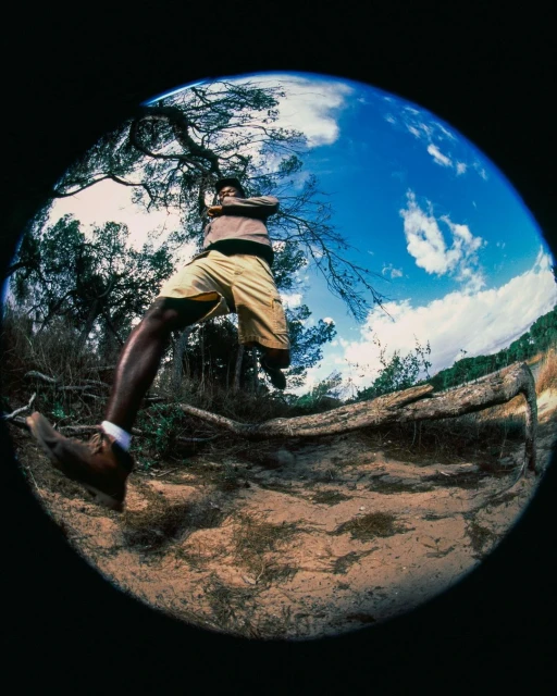 Man in mid-air wearing tan shorts and brown shoes. Fisheye lens shows trees, sky, and clouds with a black background around the circular image.