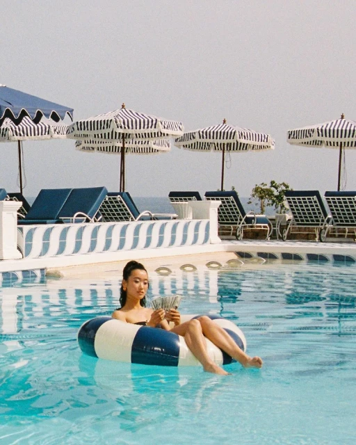 Woman in a blue and white striped inflatable ring floats in a pool holding cash near lounge chairs with striped umbrellas.