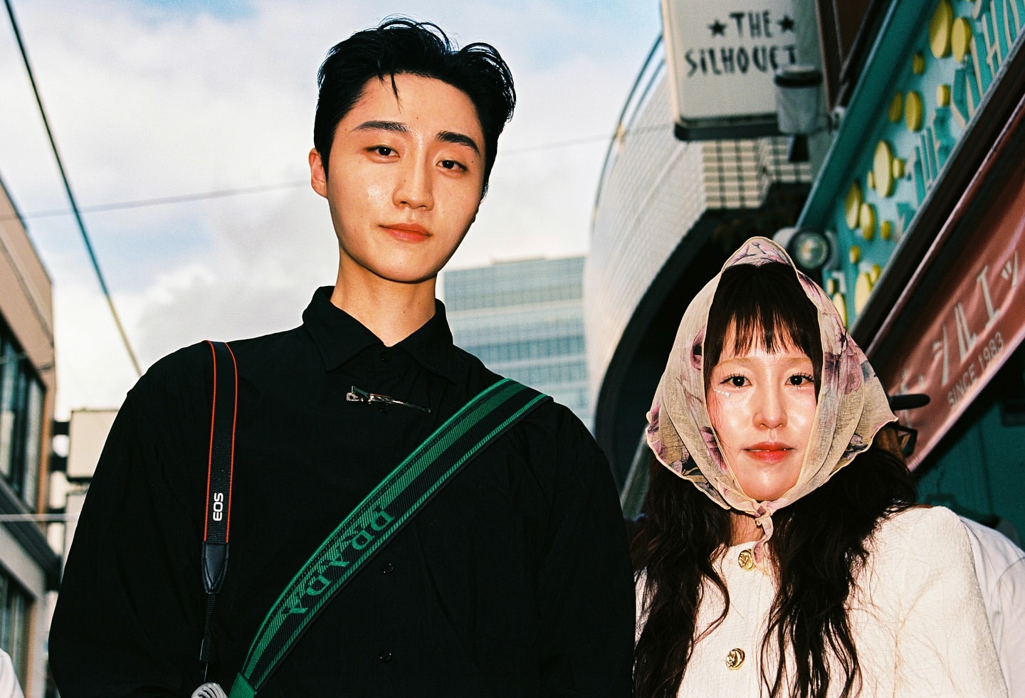 Portrait of an Asian couple outdoors in front of a building with signage, the male wears a Prada camera strap, the female a floral headscarf.