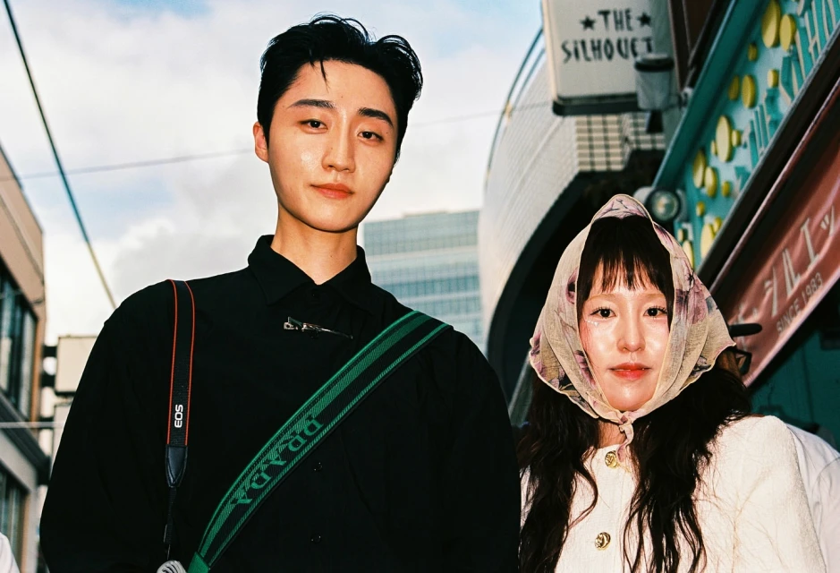 Portrait of an Asian couple outdoors in front of a building with signage, the male wears a Prada camera strap, the female a floral headscarf.