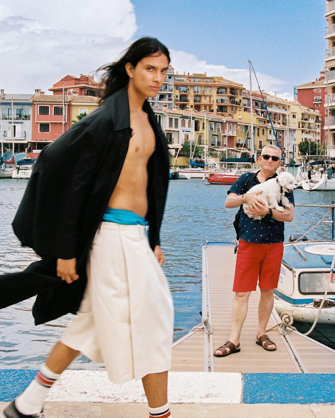 A stylish man in an open black coat and cream culottes is walking near an older man holding a white dog in a waterfront scene.
