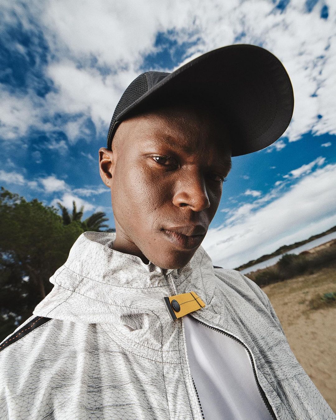 Man in athletic jacket and cap stands against a blue sky with clouds and shoreline in an eye-level shot, focusing on fashion and outdoor style.