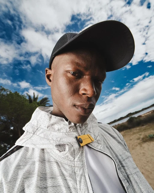 Man in athletic jacket and cap stands against a blue sky with clouds and shoreline in an eye-level shot, focusing on fashion and outdoor style.