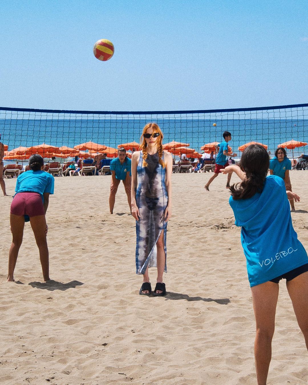 A group of women playing volleyball on a sunny beach with a volleyball net, blue sky, and orange umbrellas in the background.