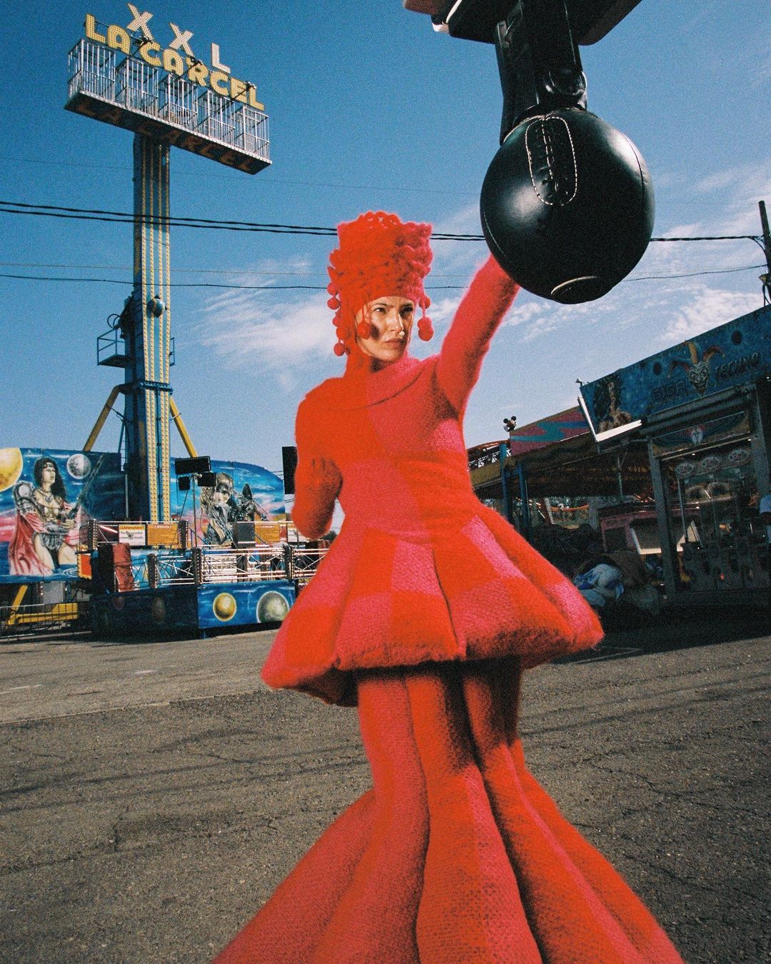 Woman in a red dress and elaborate headpiece stands in front of a carnival ride with a black punching bag prop, photographed at eye-level.