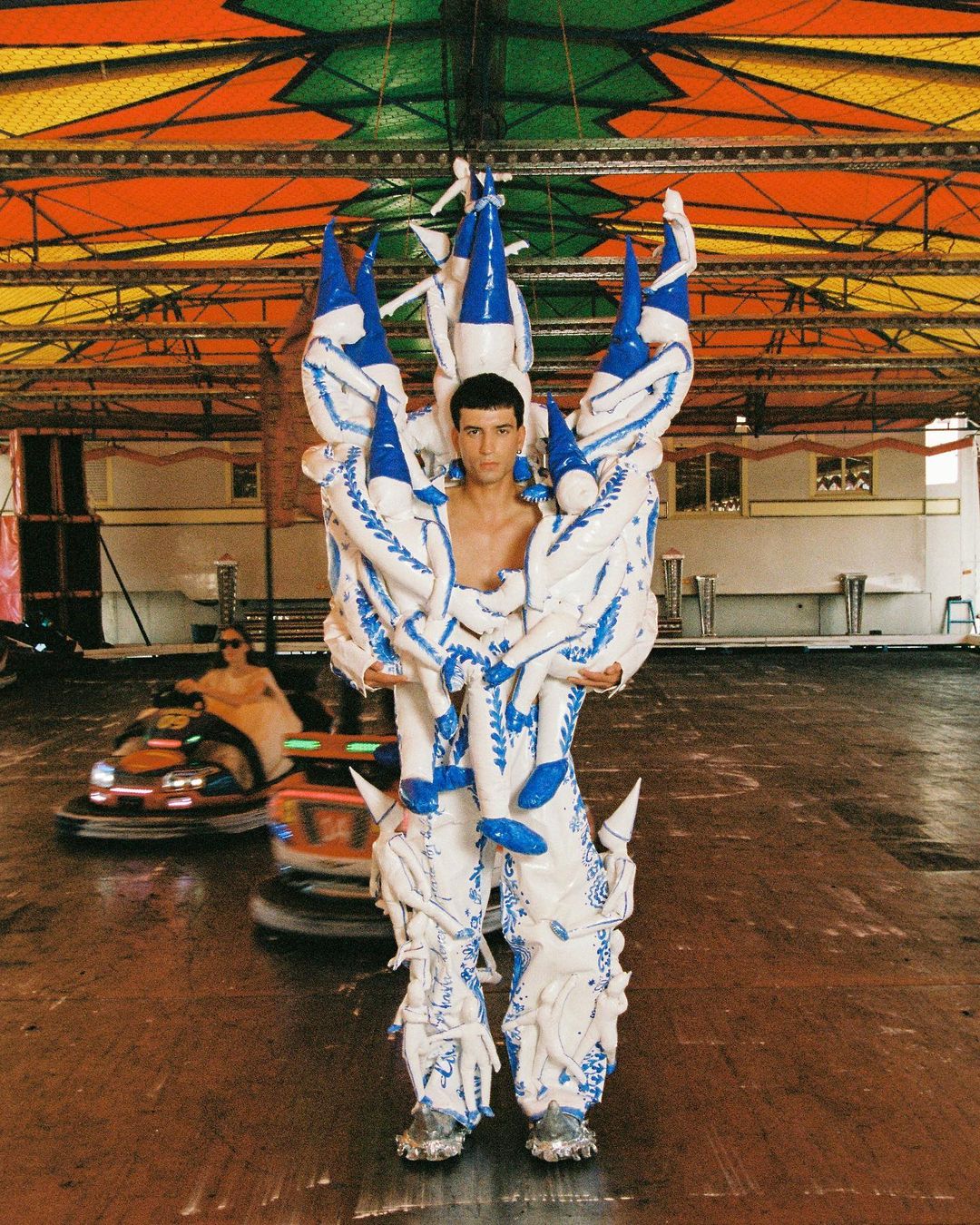 A person stands inside a carnival structure wearing an elaborate white and blue costume, with bumper cars in the background.