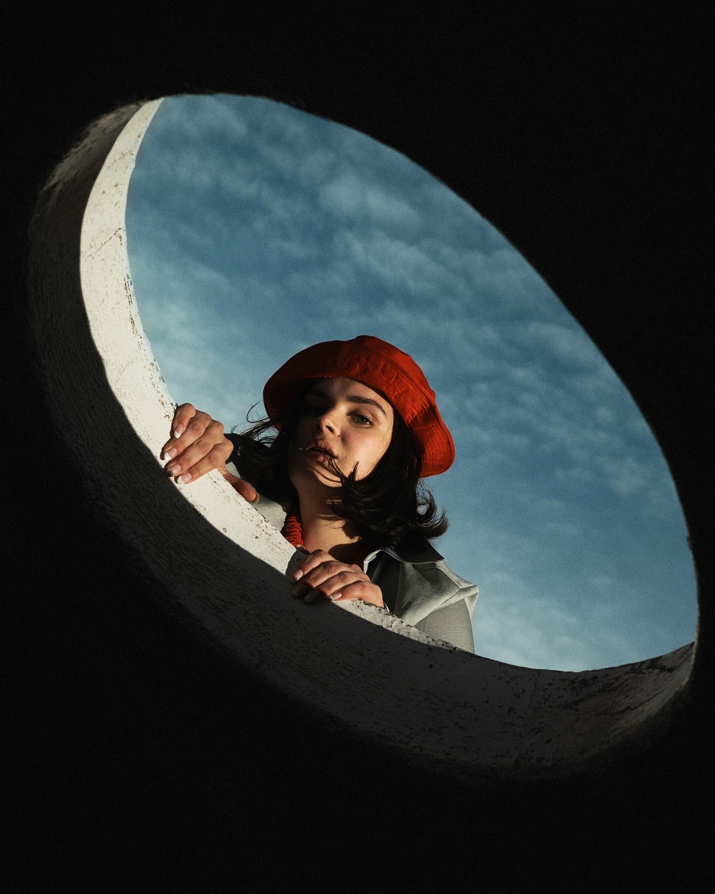Low-angle view of a woman in a red beret looking through a concrete circle against a blue sky with white clouds, natural light.