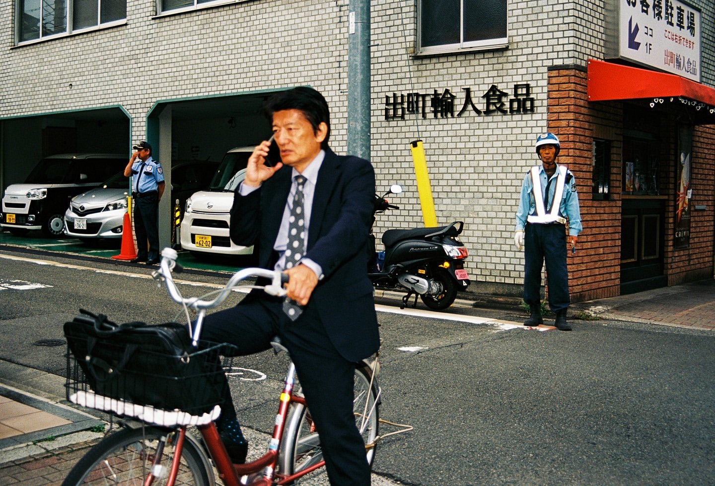 An Asian businessman in a suit rides a red bicycle while talking on his cell phone on a city street with a guard standing nearby.