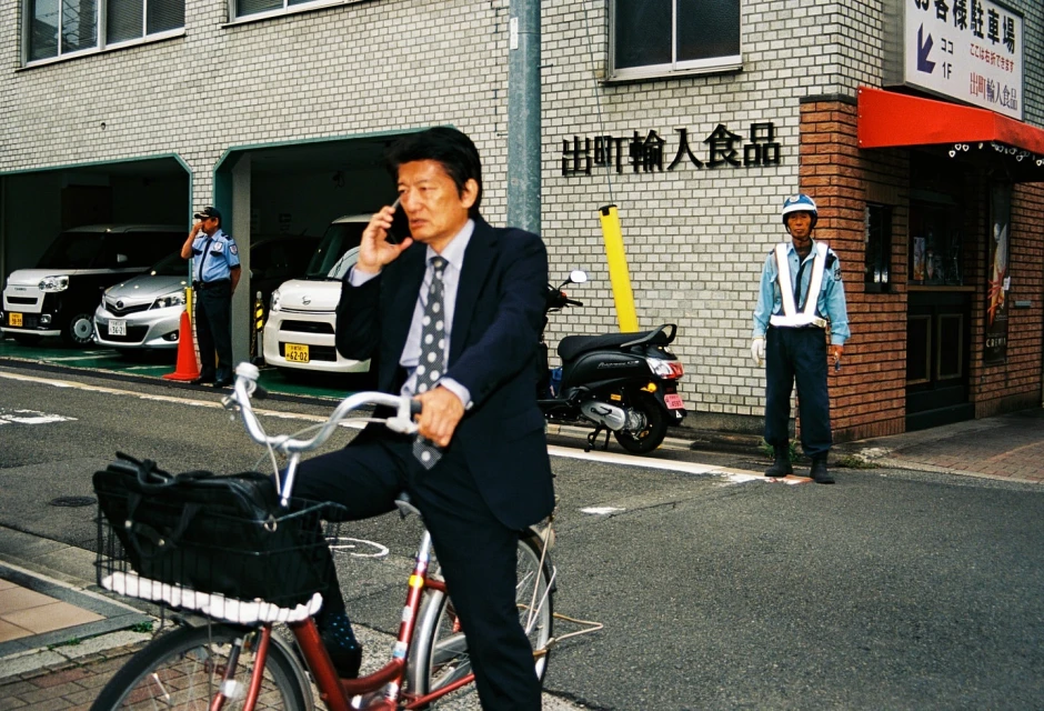 An Asian businessman in a suit rides a red bicycle while talking on his cell phone on a city street with a guard standing nearby.